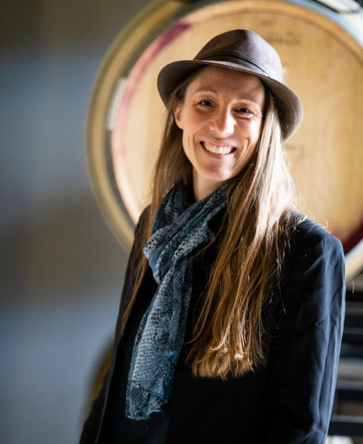 Camille Gaïo smiling in a hat and scarf in front of a wine barrel